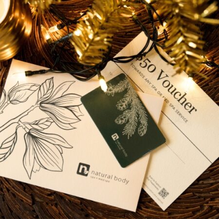 Natural Body Spa holiday gift card and $50 holiday voucher displayed on table with gold holiday tree
