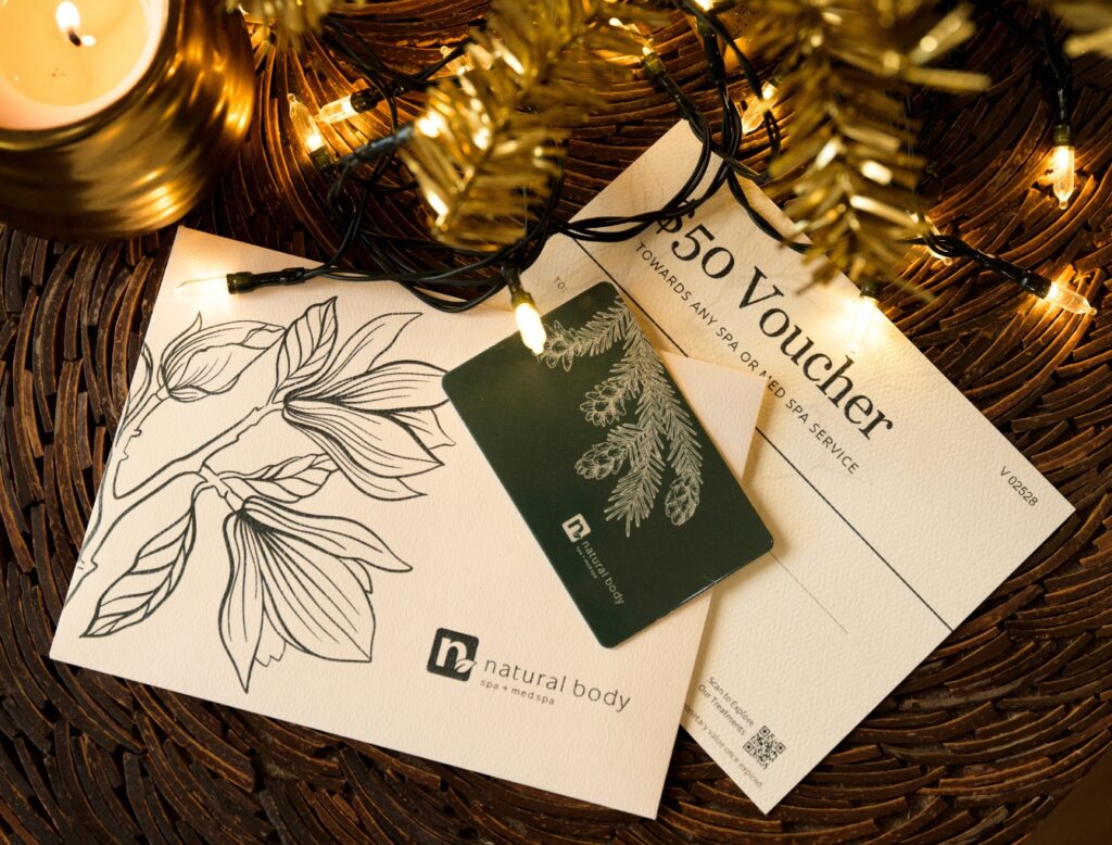 Natuarl Body Gift Card nd $50 Holiday Voucher laying on table with a gold candle and Christmas lights at Natural Body Spa + Med Spa in Atlanta, GA.